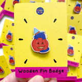 A wooden pin badge featuring a purple cat wearing a blue witches hat and standing in front of a lit halloween pumpkin is on a yellow and pink cardboard display backer, with the words Wooden Pin Badge at the bottom