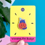 A wooden pin badge featuring a purple cat wearing a blue witches hat and standing in front of a lit halloween pumpkin is shown on a yellow and pink backing card against a brightly coloured wall